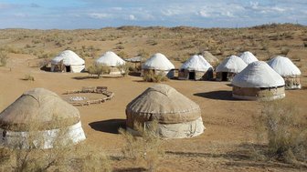 Eine Gruppe von Jurten steht in einer trockenen, sandigen Landschaft unter einem klaren Himmel mit wenigen Wolken.