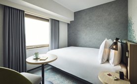 Ein modernes Hotelzimmer mit einem großen Bett, einem kleinen Tisch und einem Fenster, das viel Tageslicht hereinlässt.