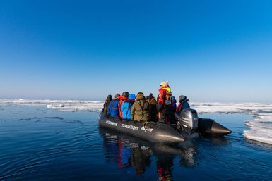 Eine Gruppe von Menschen in bunten Jacken steht auf einem Schlauchboot, umgeben von Eis und klarem blauen Himmel.