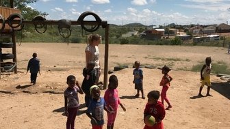 Eine Gruppe von Kindern spielt fröhlich im Freien, während eine Erwachsene sie beaufsichtigt, unter einem strahlend blauen Himmel.