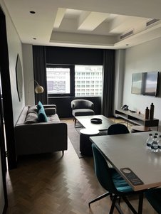 Ein stilvoller Wohnbereich mit einem grauen Sofa, einem runden Tisch und einem großen Fenster, das auf eine Stadtansicht blickt.