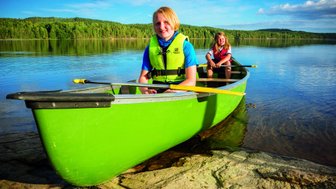 Zwei Kinder sitzen in einem grünen Kanu auf einem ruhigen See, umgeben von üppigem Wald und klarem Himmel.