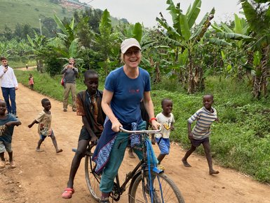 Eine Frau fährt auf einem Fahrrad, während Kinder fröhlich auf einem staubigen Weg hinterherlaufen und lachen.