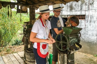 Eine Frau und ein Mann mahlen Kaffeebohnen an einer alten Mühle unter einem Strohdach in einer ländlichen Umgebung.