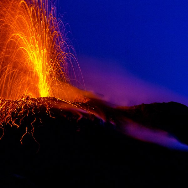 Ein aktiver Vulkan spuckt glühende Lava und Asche in den Nachthimmel, während orangefarbene Funken aufsteigen.