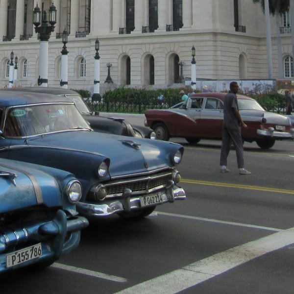 Blaue Oldtimer parken auf einer belebten Straße vor einem historischen Gebäude in Havanna, Kuba, während Passanten vorbeigehen.