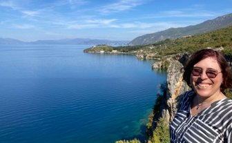 Eine lächelnde Frau steht am Rand einer Klippe mit Blick auf den ruhigen Ohridsee und die umliegenden Berge.