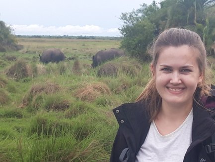 Eine lächelnde Frau steht in einer grünen Landschaft, während im Hintergrund zwei Nashörner grasen.