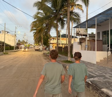Zwei Jungen gehen entspannt eine sandige Straße entlang, umgeben von Palmen und kleinen Geschäften im Hintergrund.
