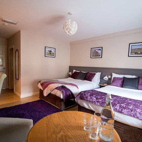 Ein gemütliches Hotelzimmer mit zwei Betten, lila Akzenten und einem runden Holztisch im Vordergrund.