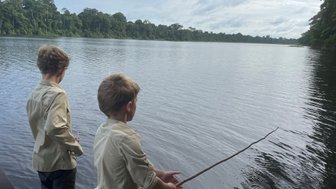 Zwei Jungen stehen am Ufer eines ruhigen Gewässers und versuchen, mit einem Stock zu fischen, umgeben von üppigem Grün.