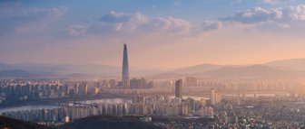 Die Skyline von Seoul bei Sonnenuntergang, mit dem markanten Lotte World Tower, umgeben von sanften Hügeln und Wolken.