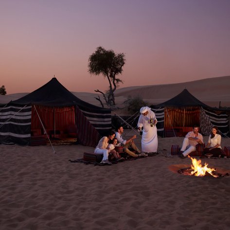 Eine Gruppe von Menschen sitzt um ein Lagerfeuer in der Wüste, während ein Beduine Tee serviert und die Dämmerung einsetzt.