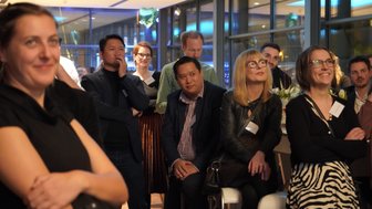 Eine Gruppe von Menschen hört aufmerksam zu, während sie in einem modernen Raum mit großen Fenstern versammelt sind.