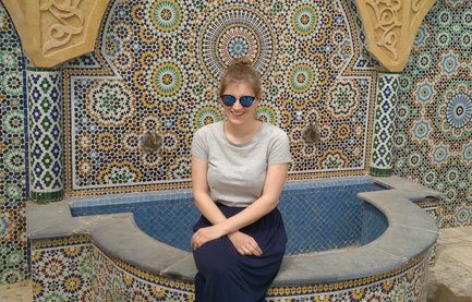 Eine junge Frau sitzt lächelnd an einem mit bunten Mosaiken verzierten Brunnen in einem marokkanischen Innenhof.