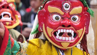 Ein Tänzer trägt eine auffällige rote Maske mit großen Augen und einem breiten Grinsen, während er in traditioneller Kleidung posiert.