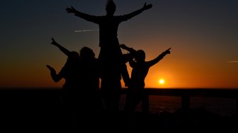 Fünf Silhouetten von Menschen, die fröhlich vor einem Sonnenuntergang posieren, während die Sonne am Horizont untergeht.