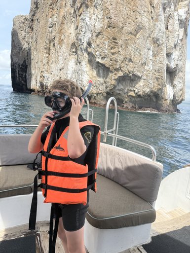 Ein Junge mit einer Schnorchelmaske und orangefarbener Schwimmweste steht auf einem Boot vor einer beeindruckenden Felswand.