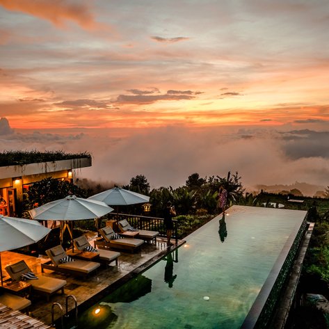 Ein atemberaubender Sonnenuntergang über einem Infinity-Pool, umgeben von üppiger Vegetation und Wolken.