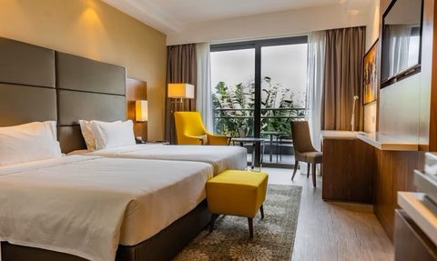 Ein modernes Hotelzimmer mit zwei Einzelbetten, einem gelben Hocker und einem Blick auf einen Balkon mit Pflanzen.