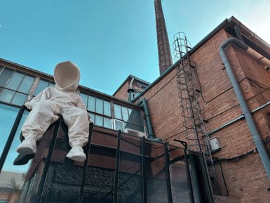 Eine große, weiße Figur sitzt auf einem Glasgeländer, umgeben von einer Backsteinmauer und einem blauen Himmel.