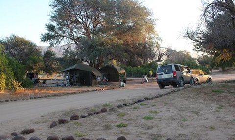 Ein Campingplatz mit einem großen Baum, einem Zelt und einem Wohnmobil, umgeben von einer ruhigen, natürlichen Umgebung.