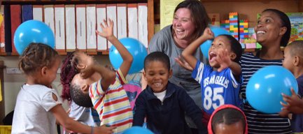 Eine Gruppe fröhlicher Kinder spielt in einem Raum mit blauen Ballons, während eine Erwachsene mit ihnen lacht.