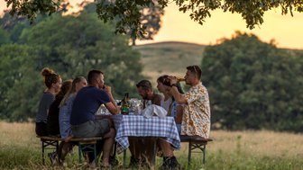 Eine Gruppe von Freunden sitzt an einem Tisch im Freien, umgeben von Natur, und genießt ein gemeinsames Essen bei Sonnenuntergang.