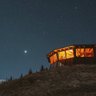 Eine beleuchtete Hütte steht auf einem Hügel, umgeben von dunklem Himmel und funkelnden Sternen.