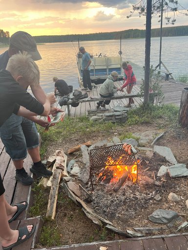 Am Ufer eines ruhigen Sees bereiten Kinder und Erwachsene gemeinsam ein Lagerfeuer vor, während die Sonne untergeht.