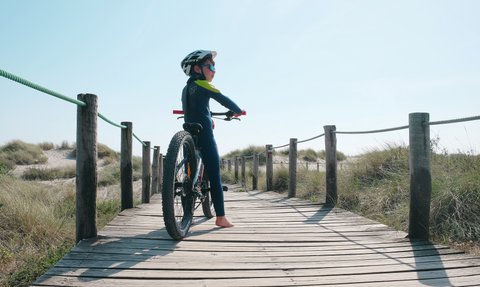 Ein Junge in einem blauen Neoprenanzug steht auf einem Holzsteg und schaut in die Ferne, während er sein Mountainbike hält.