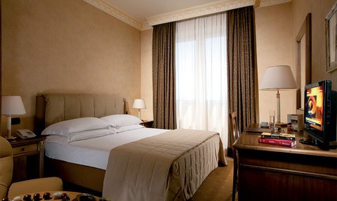 Ein elegantes Hotelzimmer mit einem großen Bett, einem Schreibtisch und einem Fernseher, umgeben von warmen Farbtönen.