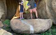Ein Mann und ein Kind stehen unter einem großen Felsen, umgeben von grünen Pflanzen und weiteren Steinen.