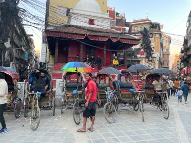 In einer belebten Straße in Kathmandu warten mehrere Rikschafahrer mit bunten Regenschirmen auf Passagiere.
