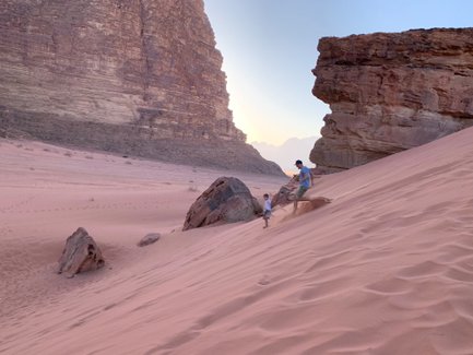 Ein Erwachsener und ein Kind spielen im weichen, rosa Sand einer Wüstenlandschaft, umgeben von hohen Felsen.