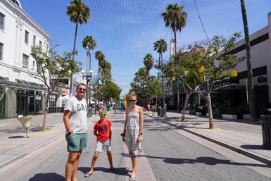 Eine Familie steht fröhlich auf der Santa Monica Promenade, umgeben von Palmen und Geschäften unter blauem Himmel.