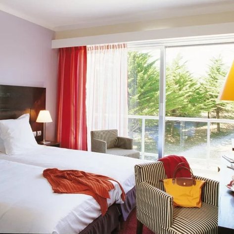 Ein helles Hotelzimmer mit zwei Betten, einem gemütlichen Sessel und einem großen Fenster, das auf einen grünen Garten blickt.