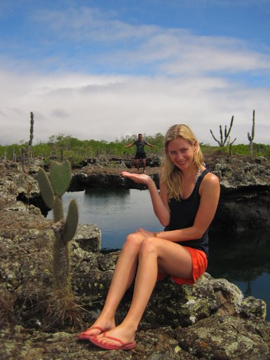 Eine Frau sitzt auf einem Felsen am Wasser, während sie einen Mann im Hintergrund mit ausgebreiteten Armen zeigt.