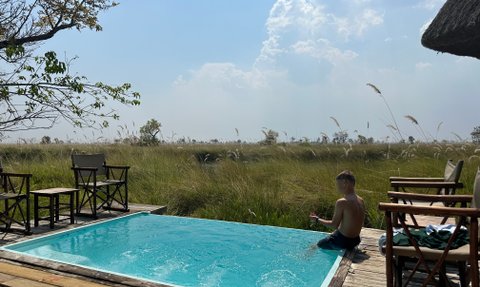 Ein Junge sitzt am Rand eines Pools und spielt mit Wasser, umgeben von hohen Gräsern und einem klaren Himmel.