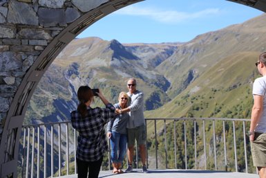 Eine Frau fotografiert ein Paar, das vor einer beeindruckenden Berglandschaft posiert, während ein weiterer Mann im Hintergrund steht.