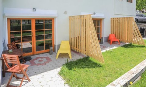 Ein sonniger Außenbereich mit Holzstühlen und bunten Sitzgelegenheiten, umgeben von grünem Gras und einem gepflegten Garten.