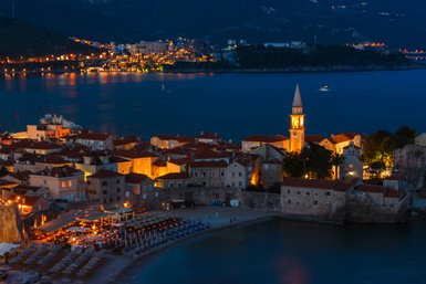 Die Küstenstadt Budva erstrahlt nachts in warmem Licht, während der Glockenturm im Vordergrund leuchtet.