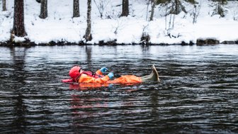 Eine Person in einem orangefarbenen Anzug schwimmt entspannt auf einem ruhigen Fluss, umgeben von schneebedeckten Bäumen.