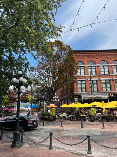 Eine belebte Straßenszene in Vancouver mit einem historischen Gebäude und Gästen, die unter gelben Sonnenschirmen sitzen.
