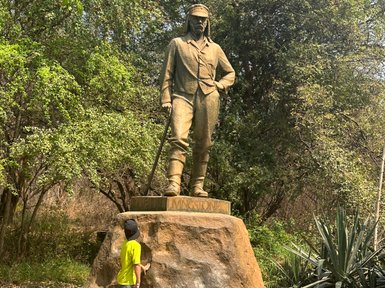 Ein Junge in einem leuchtend grünen T-Shirt steht vor einer Statue von David Livingstone, umgeben von üppigem Grün.