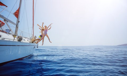 Drei fröhliche Personen springen von einem Segelboot ins glitzernde blaue Wasser, umgeben von strahlendem Sonnenschein.