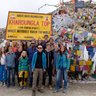 Eine Gruppe von Reisenden posiert fröhlich vor dem Khardungla-Pass-Schild, umgeben von bunten Gebetsfahnen.