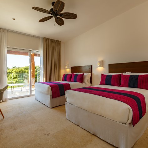 Ein helles Hotelzimmer mit zwei Betten, dekoriert mit bunten Kissen und Blick auf eine grüne Landschaft durch große Fenster.