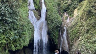Ein Wasserfall stürzt in einen klaren Pool, umgeben von üppigem Grün und Felsen, während zwei Personen die Szenerie genießen.