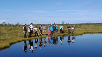 Eine Gruppe von Menschen steht am Ufer eines ruhigen Gewässers, umgeben von einer weitläufigen, grünen Moorlandschaft.